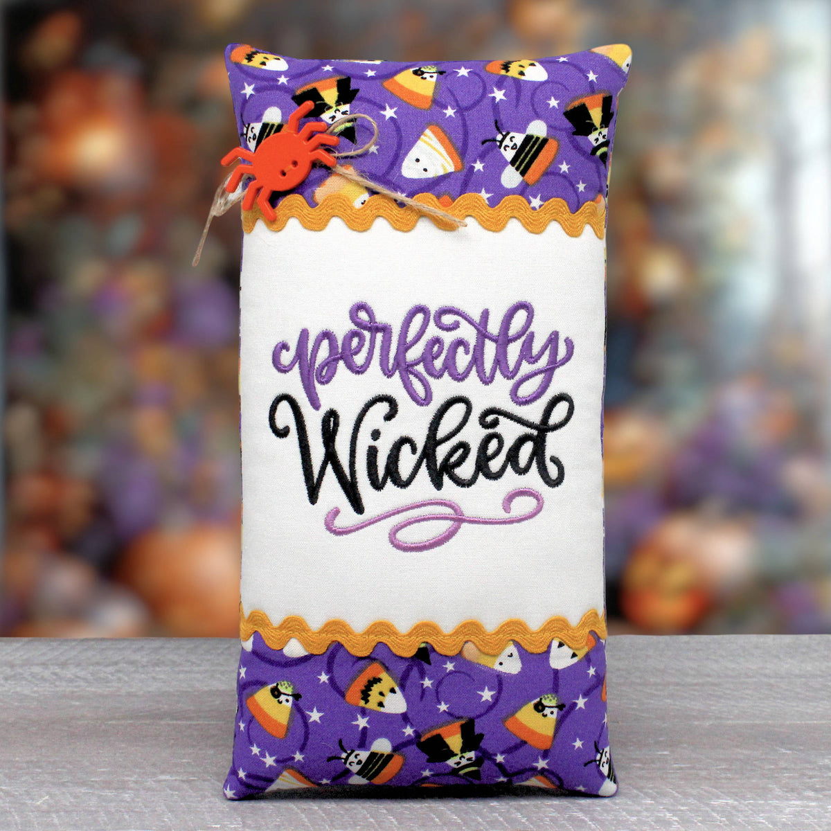 A rectangular decorative pillow with a Halloween theme, featuring the embroidered text 'perfectly Wicked' in purple and black on a white center panel. The top and bottom borders are purple with embroidered candy corn, ghosts, and stars.