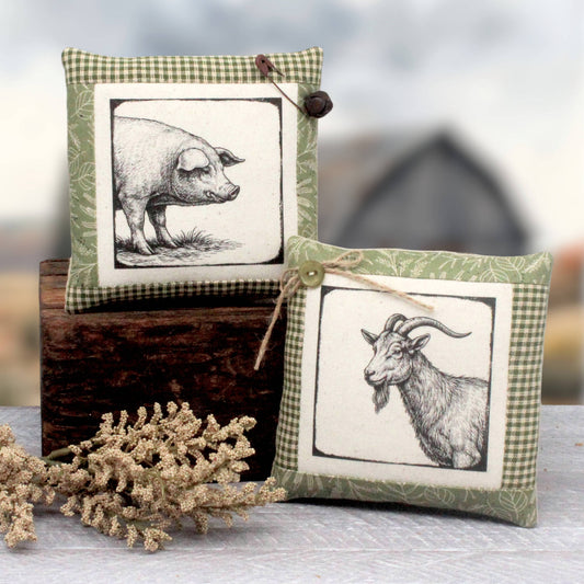 Two decorative square fabric pillows with black-and-white animal illustrations, styled on a rustic wooden block with dried flowers in the foreground against a softly blurred outdoor background.