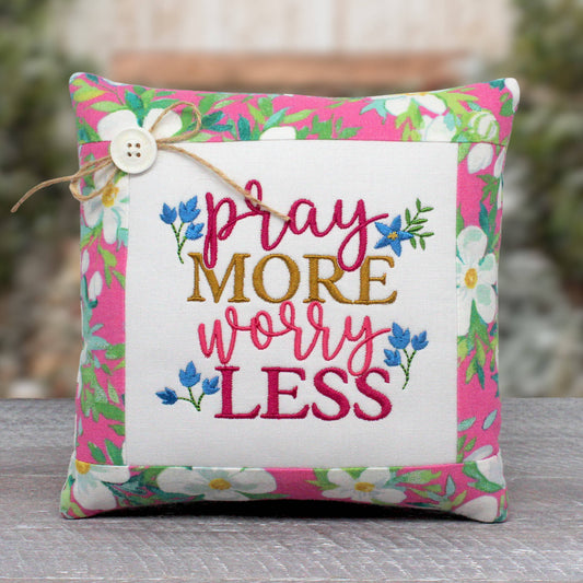 Small accent pillow or bowl filler is machine-embroidered with the phrase 'pray more worry less.' It's trimmed with a fabric that has white blossoms and green leaves on a pink background.