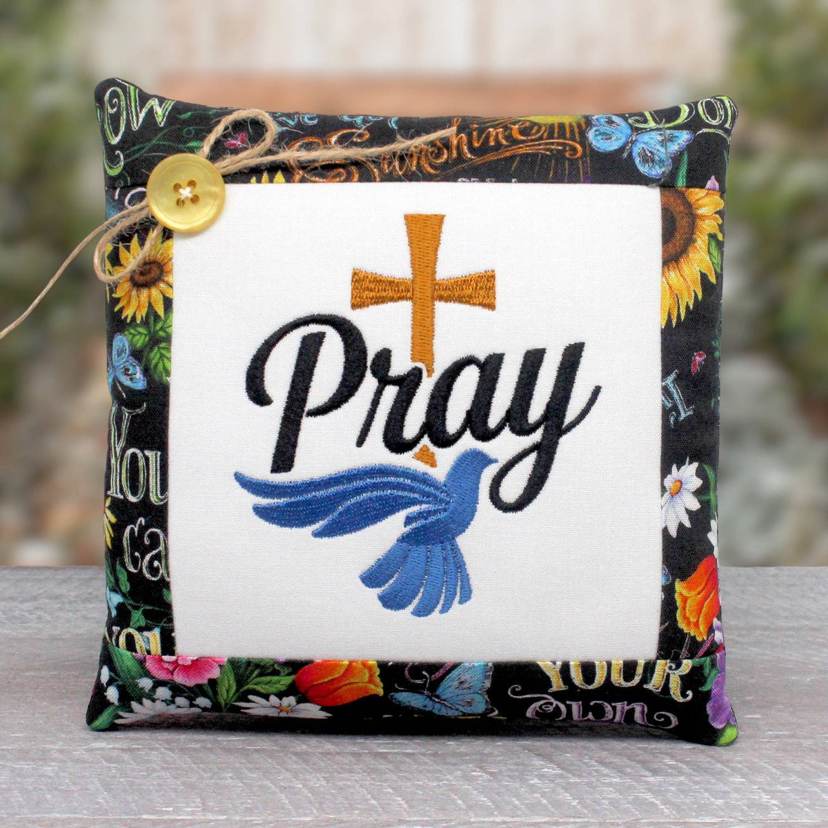 Small pillow tuck or bowl filler embroidered with the word 'pray' alongside a Christian cross and blue dove design, trimmed with a print fabric with flowers on a black background