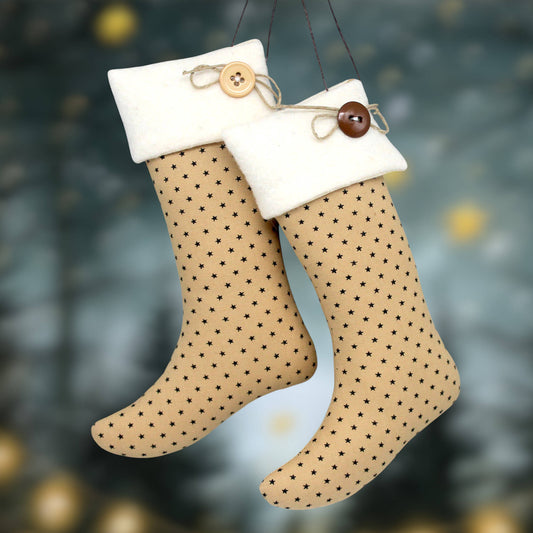 Two hanging Christmas stockings with tan fabric bodies, small black star patterns, and white fleece cuffs. One cuff has a light wood button with twine, and the other has a darker brown wooden button with twine, set against a blurred festive background.