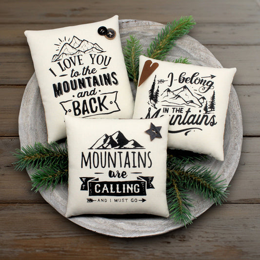 Three pillows with mountain-themed sayings on a wooden surface with pine branches.