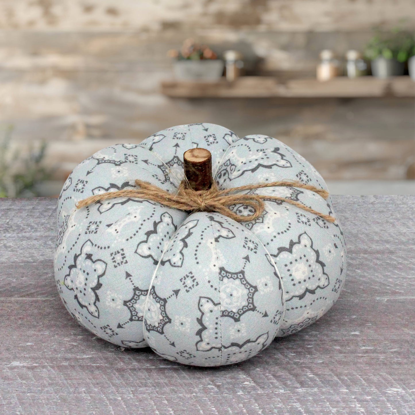 A decorative fabric pumpkin in a pale blue and gray bandana pattern, tied with twine and topped with a small wooden stem, set on a rustic gray surface with a blurred stone wall background.