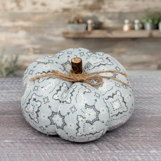A decorative fabric pumpkin in a pale blue and gray bandana pattern, tied with twine and topped with a small wooden stem, set on a rustic gray surface with a blurred stone wall background.