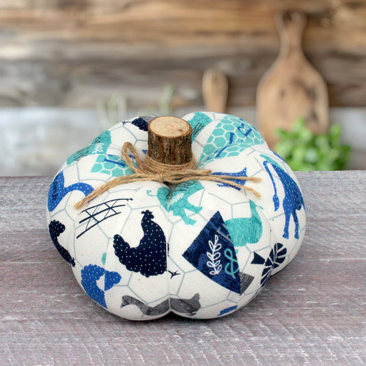 A fabric decorative pumpkin with a rustic wood stem, featuring a patchwork print of blue, teal, and gray farm and autumn motifs on an off-white background, tied with a piece of twine.