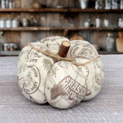 A decorative faux pumpkin with a rustic off-white fabric body printed with vintage farmers market design and a natural wood stem, tied with twine on a gray tabletop with a blurred rustic kitchen background.