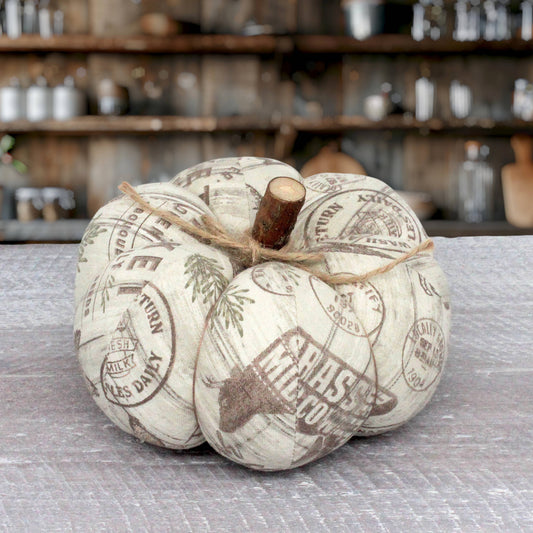 A decorative faux pumpkin with a rustic off-white fabric body printed with vintage farmers market design and a natural wood stem, tied with twine on a gray tabletop with a blurred rustic kitchen background.