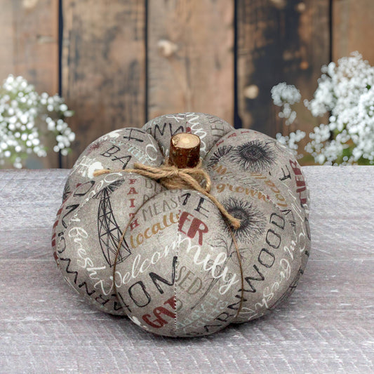 A decorative faux pumpkin with a rustic gray and beige printed pattern featuring farmhouse-themed words and illustrations, tied with a natural jute twine bow and topped with a small wooden stem, set on a gray tabletop with a blurred rustic wood background and white flowers.