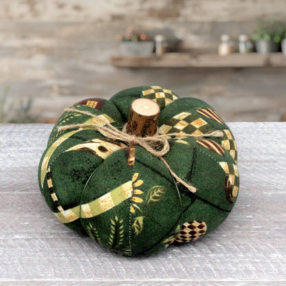 A fabric pumpkin decoration made from dark green fabric with autumn farmhouse patterns, tied with twine and topped with a small wooden stem, set on a gray tabletop with a blurred rustic background.