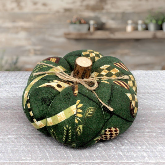 A fabric pumpkin decoration made from dark green fabric with autumn farmhouse patterns, tied with twine and topped with a small wooden stem, set on a gray tabletop with a blurred rustic background.