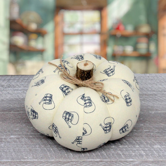 A fabric decorative pumpkin in cream with a navy blue print of hanging watering cans, tied with a rustic jute twine bow and a small wood slice stem, sitting on a gray wooden surface with a blurred background.