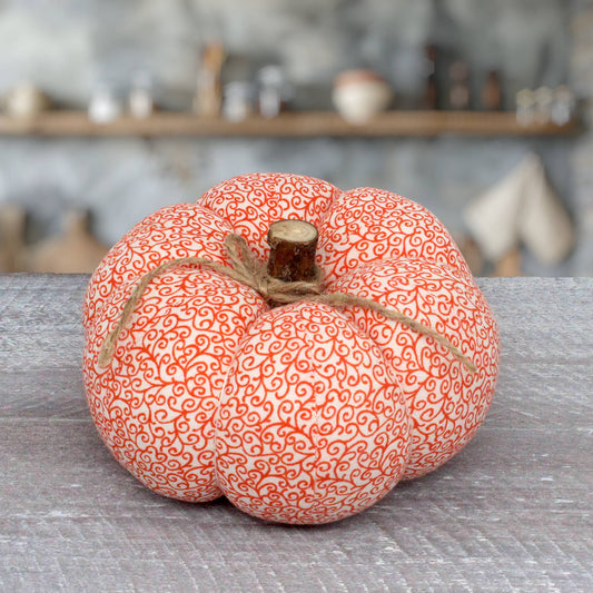 A decorative fabric pumpkin in orange and white with a swirling pattern, tied with a rustic jute twine bow and featuring a faux wood stem, set on a gray textured tabletop with a blurred rustic background.