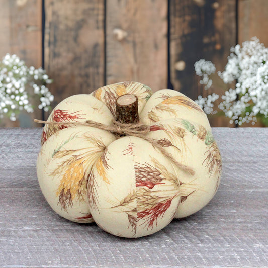 A decorative fabric pumpkin with a cream base and a wheat-like botanical print in muted red, yellow, and brown tones, tied with twine and set on a gray surface against a rustic wooden background.