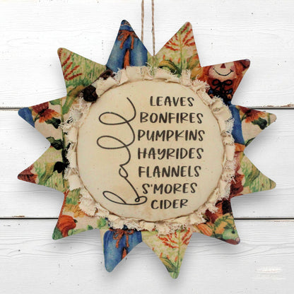 A rustic fabric sunflower wall hanging with a central cream circle that reads 'LEAVES BONFIRES PUMPKINS HAYRIDES FLANNELS S'MORES CIDER' in black script and print. The sun has multiple star points decorated with multicolored patterned fabric, including blue, green, orange, and floral designs, and is shown hanging against a white wood plank background.