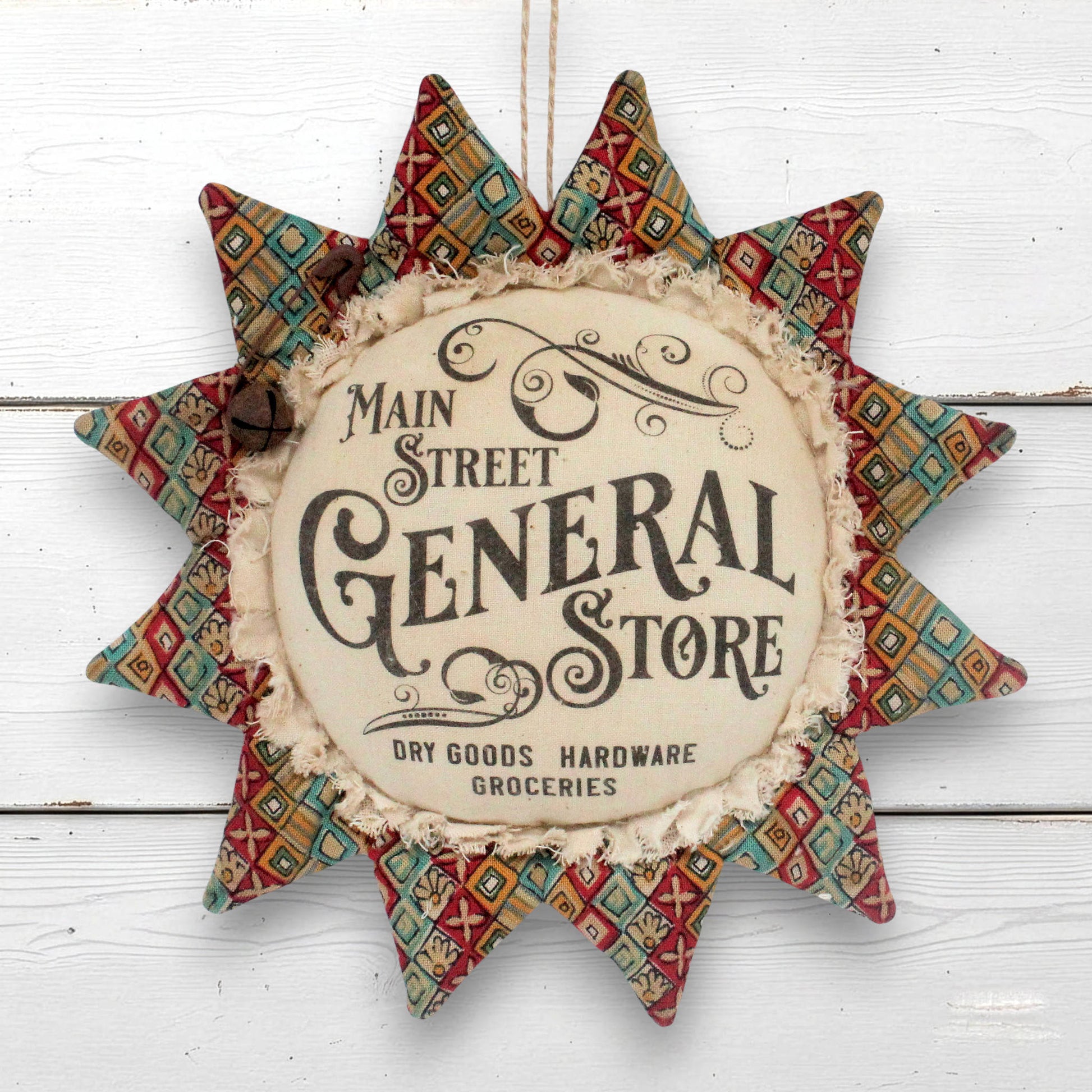A decorative fabric sunflower-shaped wall hanging with a vintage-style sign in the center reading 'Main Street General Store.' The star has patterned red, teal, beige, and brown fabric points and is photographed against a white painted wood background.