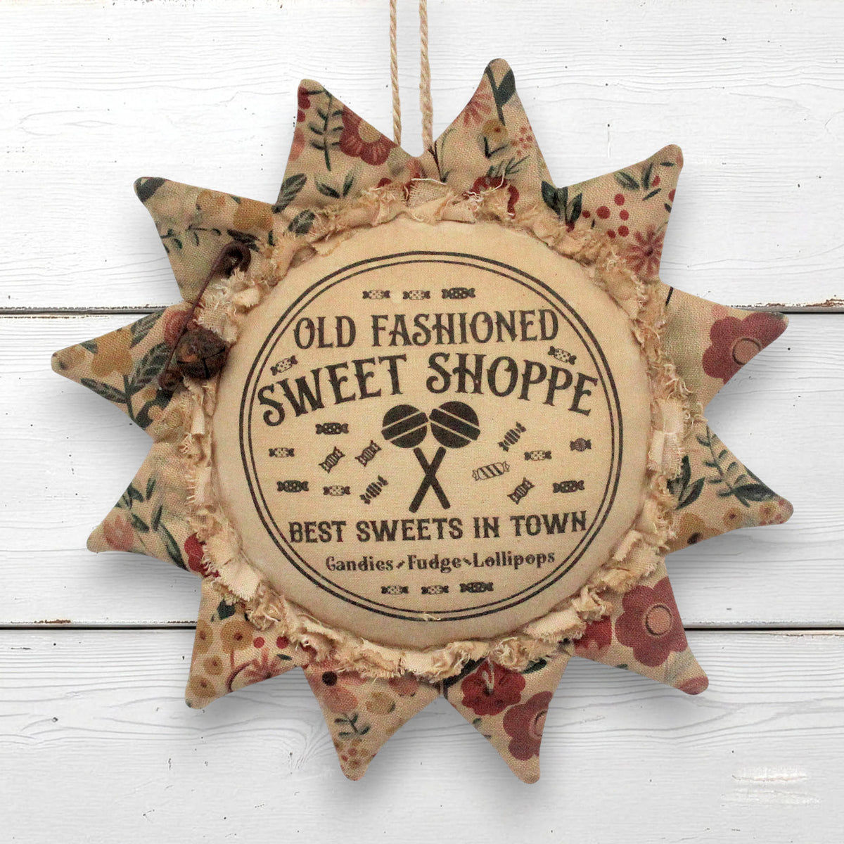 A decorative fabric sunflower-shaped ornament hanging on a white wooden wall. The center reads “Old Fashioned Sweet Shoppe” with “Best Sweets in Town” and small illustrations of lollipops, surrounded by floral-patterned star points.