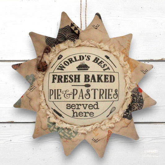 A sunflower-shaped fabric wall hanging with a rustic patchwork design and a central circular label that reads 'World's Best Fresh Baked Pie & Pastries served here,' shown against a white wood plank background.
