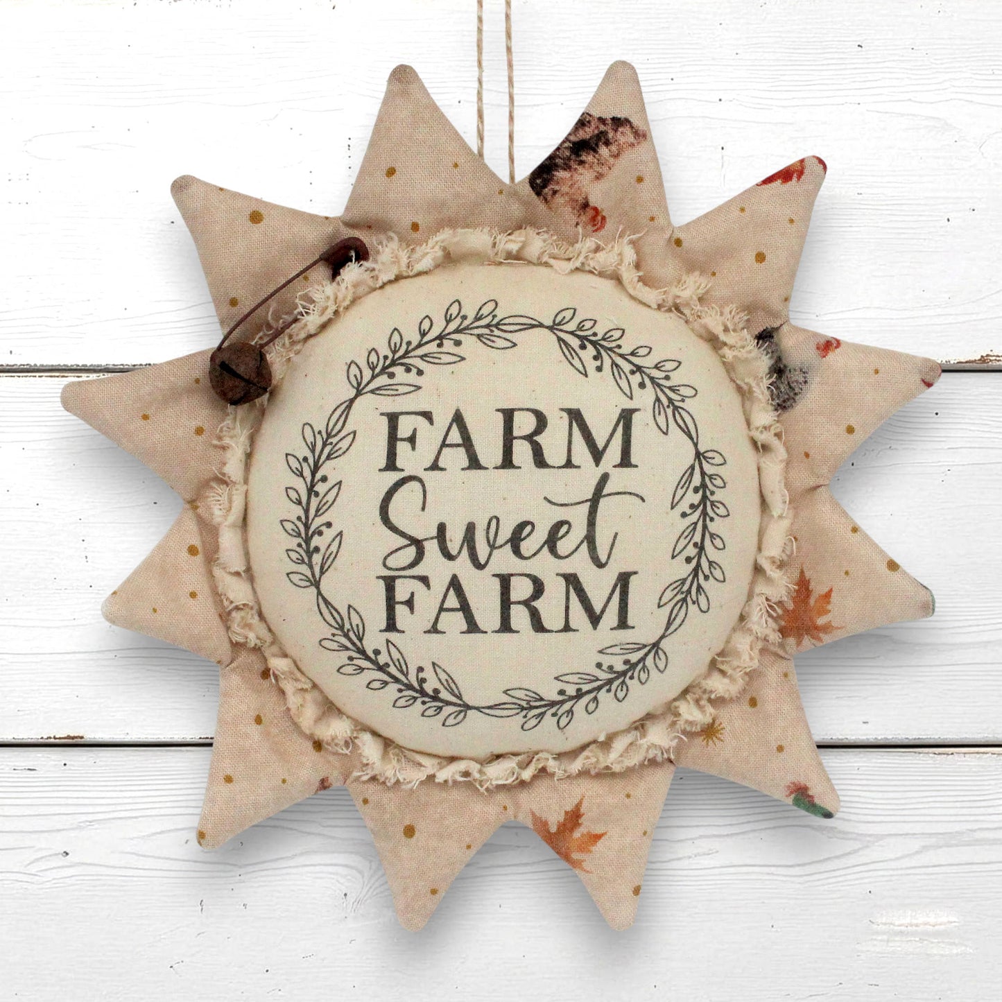 A fabric sunflower hanging ornament with the words “FARM Sweet FARM” in the center, surrounded by a leafy wreath design. The ornament has beige fabric rays with small gold speckles and a few orange leaf accents, and is photographed against a white wood plank background.