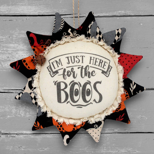 A fabric sunflower ornament featuring the text 'I'M JUST HERE for the BOOS' in black lettering. The center is cream-colored fabric with a frayed edge, surrounded by multiple star-shaped points decorated with various black, orange, red, and gray patterned fabrics.