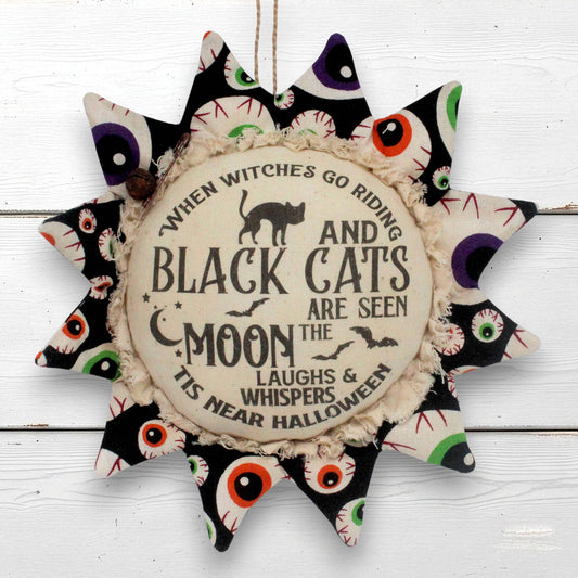 A sunflower-shaped fabric Halloween wall hanging with a central round plaque that reads, 'When witches go riding and black cats are seen the moon the laughs & whispers 'tis near Halloween.' The outer points feature black fabric with colorful embroidered or printed eyes in purple, green, red, and white.