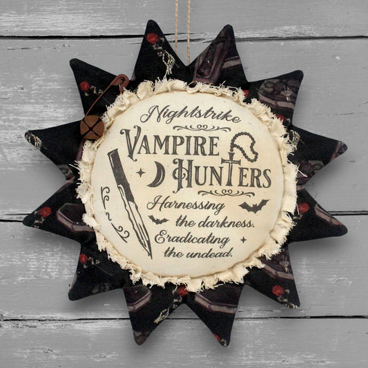 A handmade primitive sunflower fabric ornament with, featuring the text 'Nightstrike Vampire Hunters' and a dagger illustration, hanging against a gray wood plank background.