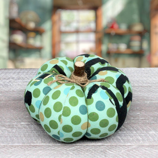 A fabric pumpkin decoration with a mint green base, black and olive green polka dots, and a small natural wood stem tied with twine, placed on a gray wooden surface with a blurred indoor background.