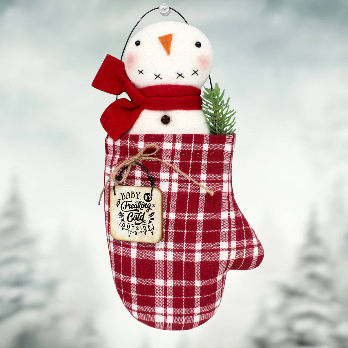 A primitive Christmas ornament with a cotton batting snowman sitting inside a red and white homespun mitten with a sprig of pine and a little wooden sign