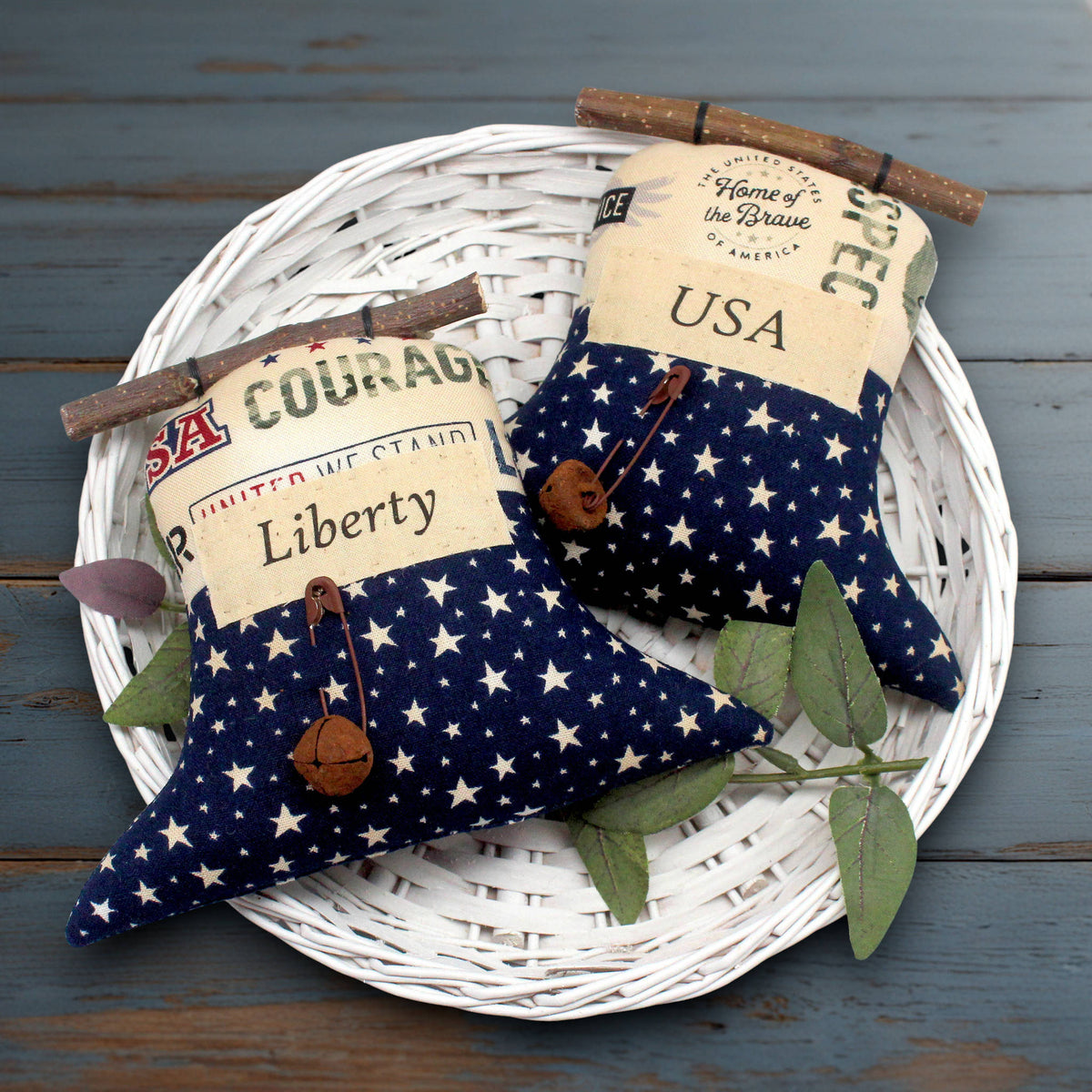 Two patriotic fabric liberty bell ornaments with a navy blue and white star pattern, decorated with vintage-style labels, wooden twigs, and small wooden bells, displayed on a white woven tray over a rustic blue wooden surface.