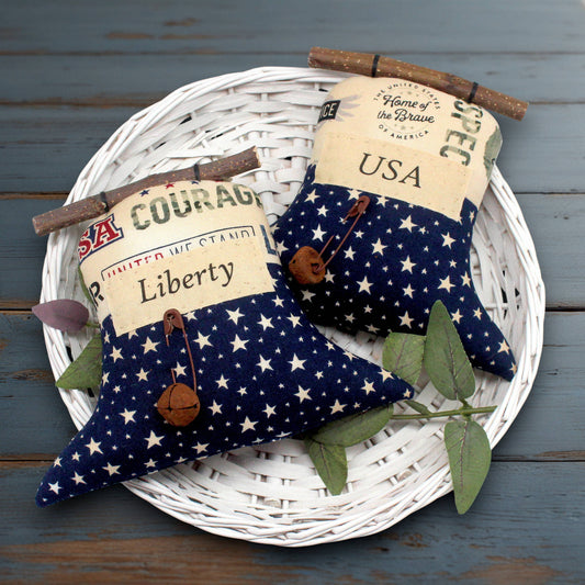 Two patriotic fabric liberty bell ornaments with a navy blue and white star pattern, decorated with vintage-style labels, wooden twigs, and small wooden bells, displayed on a white woven tray over a rustic blue wooden surface.