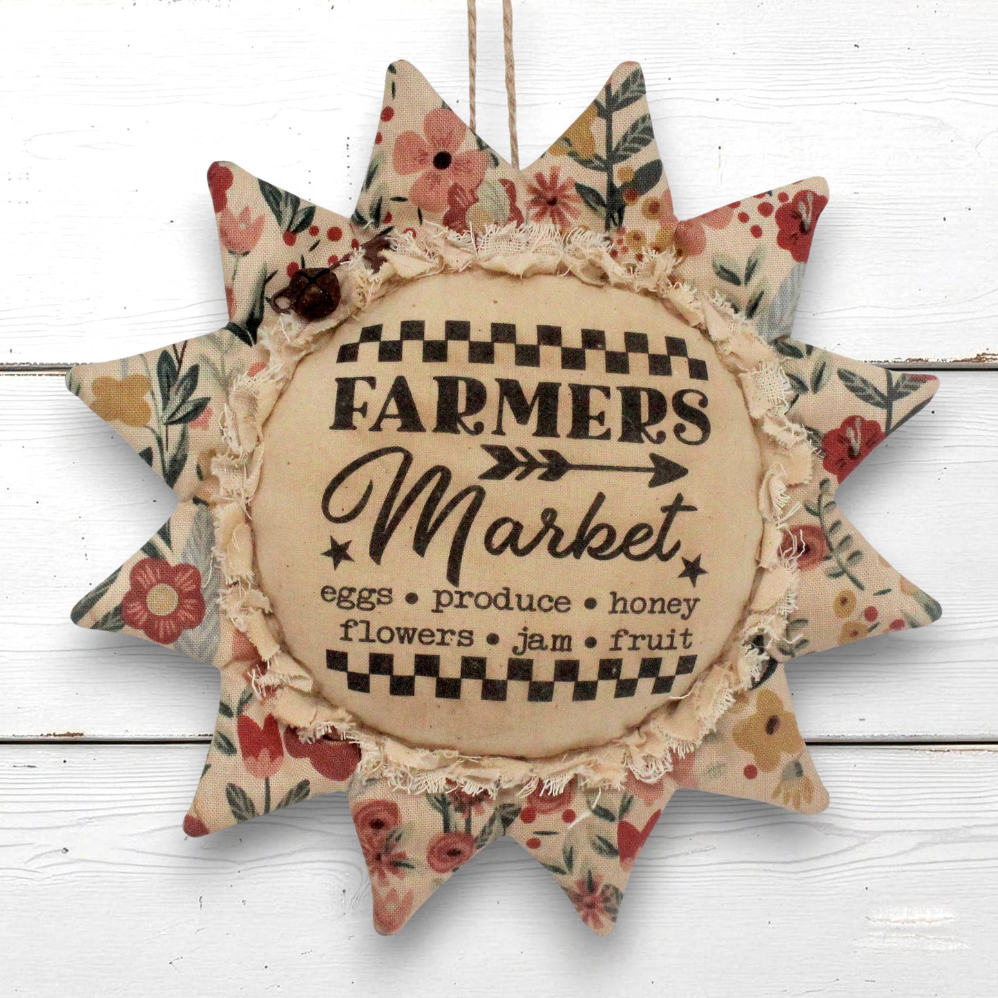 A rustic fabric sunflower ornament with floral-patterned points and a central beige circle reading “Farmers Market,” hanging against a white wood plank background.