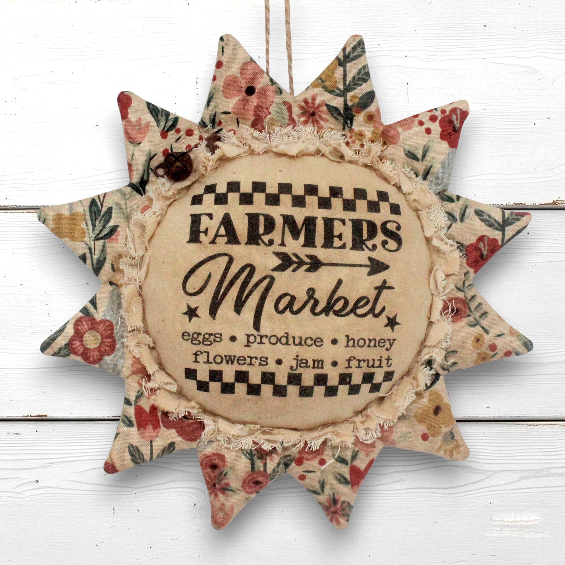 A rustic fabric sunflower ornament with floral-patterned points and a central beige circle reading “Farmers Market,” hanging against a white wood plank background.