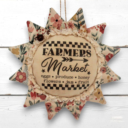 A rustic fabric sunflower ornament with floral-patterned points and a central beige circle reading “Farmers Market,” hanging against a white wood plank background.