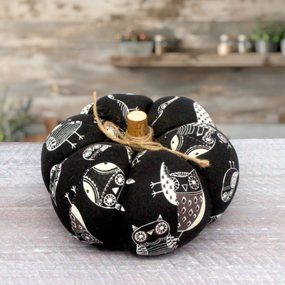 A fabric pumpkin decoration with a black background and white owl pattern, tied with natural jute twine and topped with a small wooden stem, set on a light gray surface with a blurred rustic background.