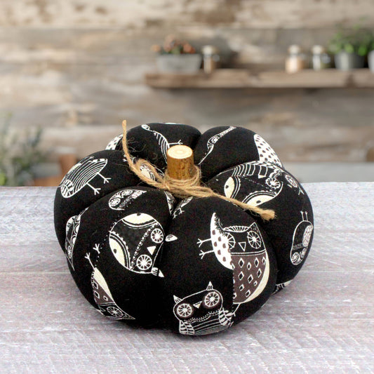 A fabric pumpkin decoration with a black background and white owl pattern, tied with natural jute twine and topped with a small wooden stem, set on a light gray surface with a blurred rustic background.