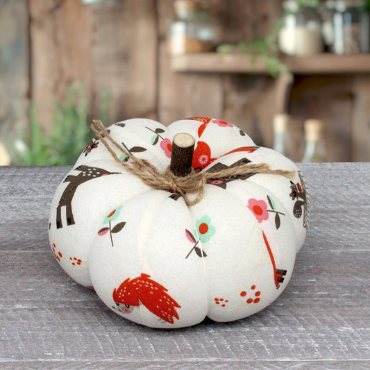 A fabric decorative pumpkin with a white base and a colorful autumn print featuring red birds, brown deer, and small flowers, tied with a rustic jute twine stem, set on a gray wooden surface with a blurred rustic background.