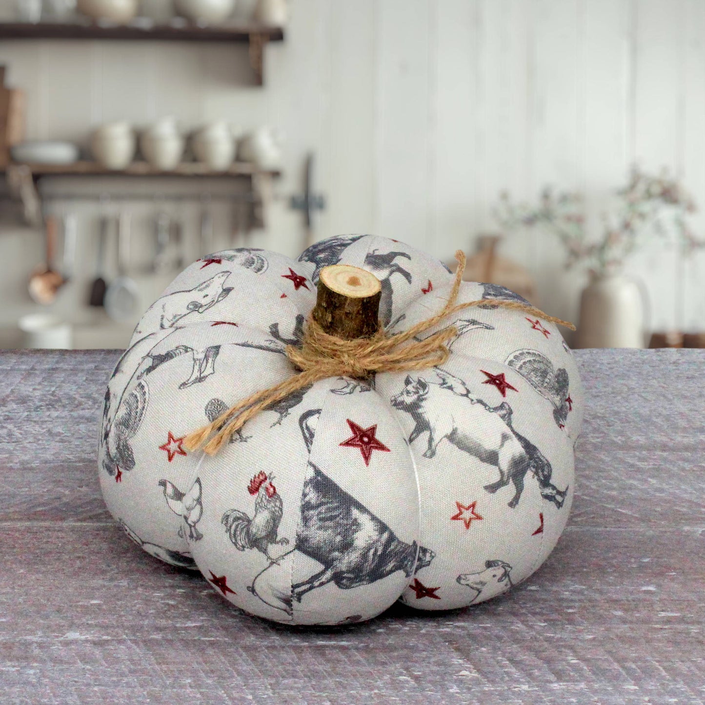 A decorative fabric pumpkin with a rustic wood stem, featuring a black and white farm animal print with red stars, tied with a jute twine bow, set on a gray wooden surface.