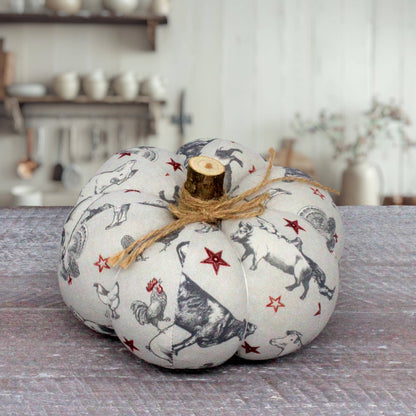 A decorative fabric pumpkin with a rustic wood stem, featuring a black and white farm animal print with red stars, tied with a jute twine bow, set on a gray wooden surface.