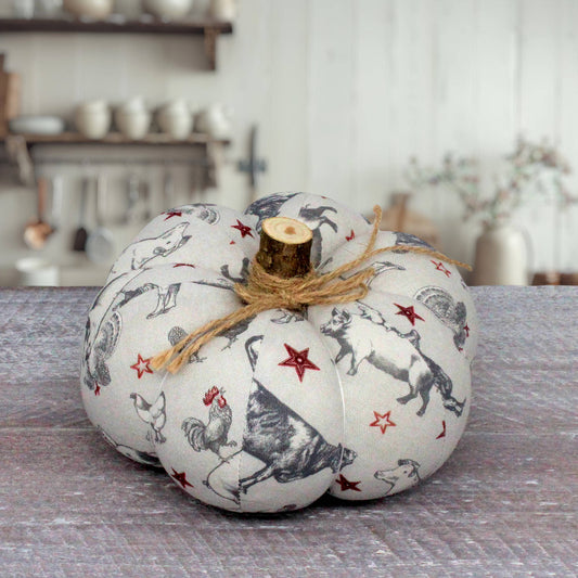 A decorative fabric pumpkin with a rustic wood stem, featuring a black and white farm animal print with red stars, tied with a jute twine bow, set on a gray wooden surface.