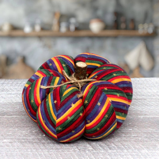 A fabric pumpkin decoration with colorful rainbow stripes and small gold stars, tied with twine and featuring a small wooden stem, displayed on a light gray textured surface.