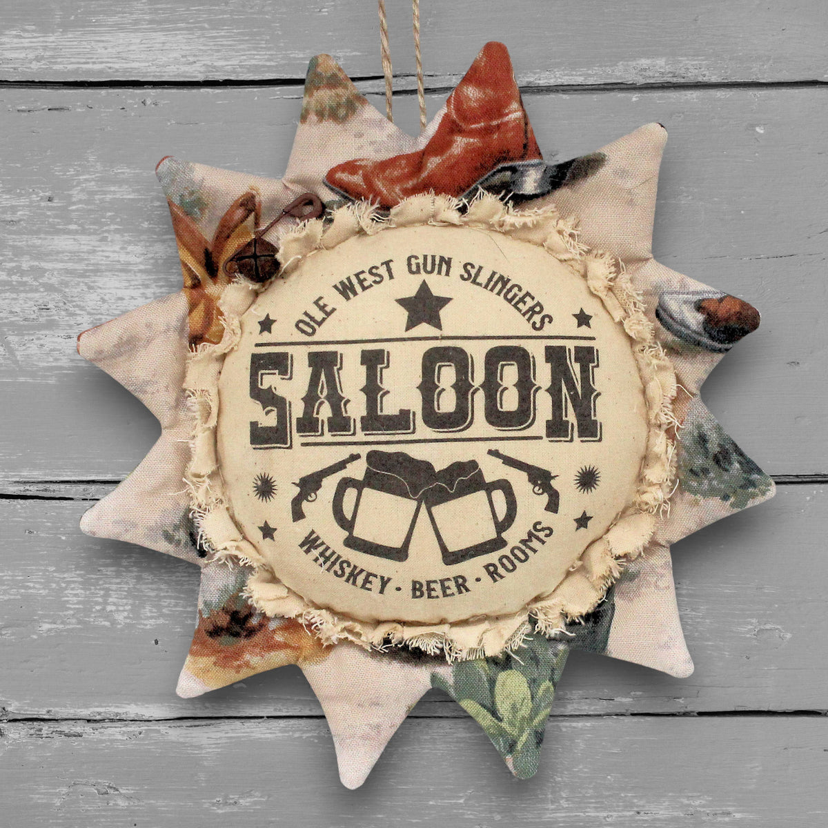 A rustic fabric sunflower-shaped ornament hanging against a gray wood plank background. The center reads “OLE WEST SALOON WHISKEY - BEER - ROOMS” with illustrations of clinking mugs and a star.