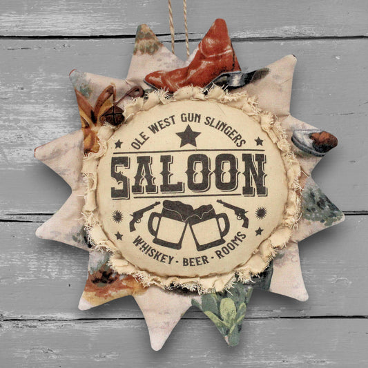 A rustic fabric sunflower-shaped ornament hanging against a gray wood plank background. The center reads “OLE WEST SALOON WHISKEY - BEER - ROOMS” with illustrations of clinking mugs and a star.