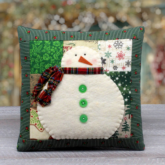 A square handmade Christmas throw pillow featuring a patchwork snowman design. The pillow has a dark green striped border, a central patchwork of green, white, and holiday-themed fabrics, a felt snowman body with three green buttons, and a red plaid scarf.