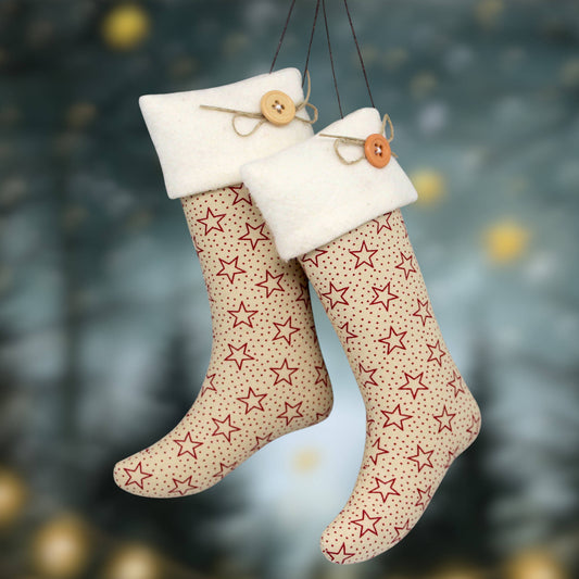 Two Christmas stockings hanging side by side against a blurred festive background. The stockings are tan with a red star pattern and have white fleece cuffs decorated with wooden buttons and twine.