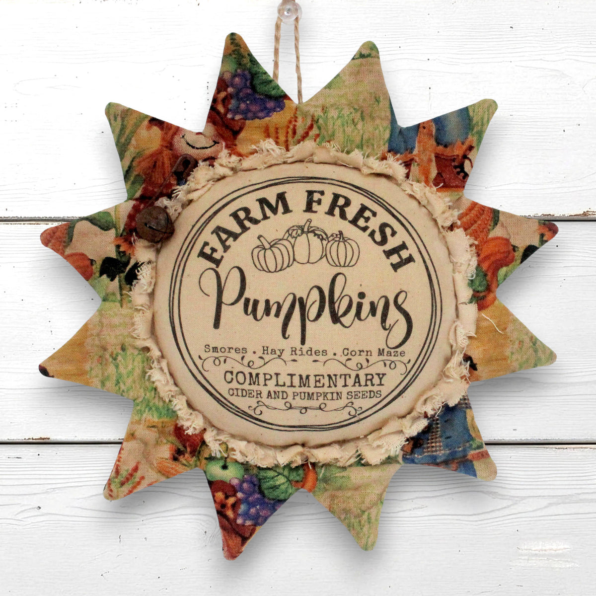 A rustic sunflower-shaped decorative hanging ornament with a central round label reading 'Farm Fresh Pumpkins' and patterned fabric points in autumn colors, shown against a white wood background.