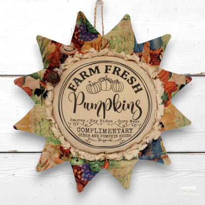 A rustic sunflower-shaped decorative hanging ornament with a central round label reading 'Farm Fresh Pumpkins' and patterned fabric points in autumn colors, shown against a white wood background.