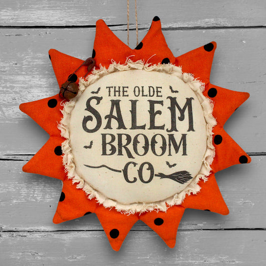 A fabric sunflower-shaped hanging ornament with orange petals, black polka dots, and a cream center reading “THE OLDE SALEM BROOM CO,” shown against a gray wood plank background.