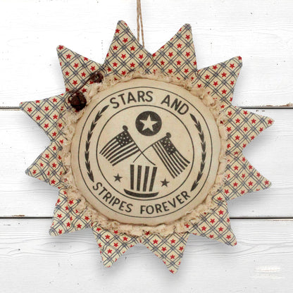 A rustic sunflower ornament hanging wall decor piece with a central round label reading “STARS AND STRIPES FOREVER” and crossed American flags, surrounded by patterned fabric rays in red, white, and blue on a white wood plank background.