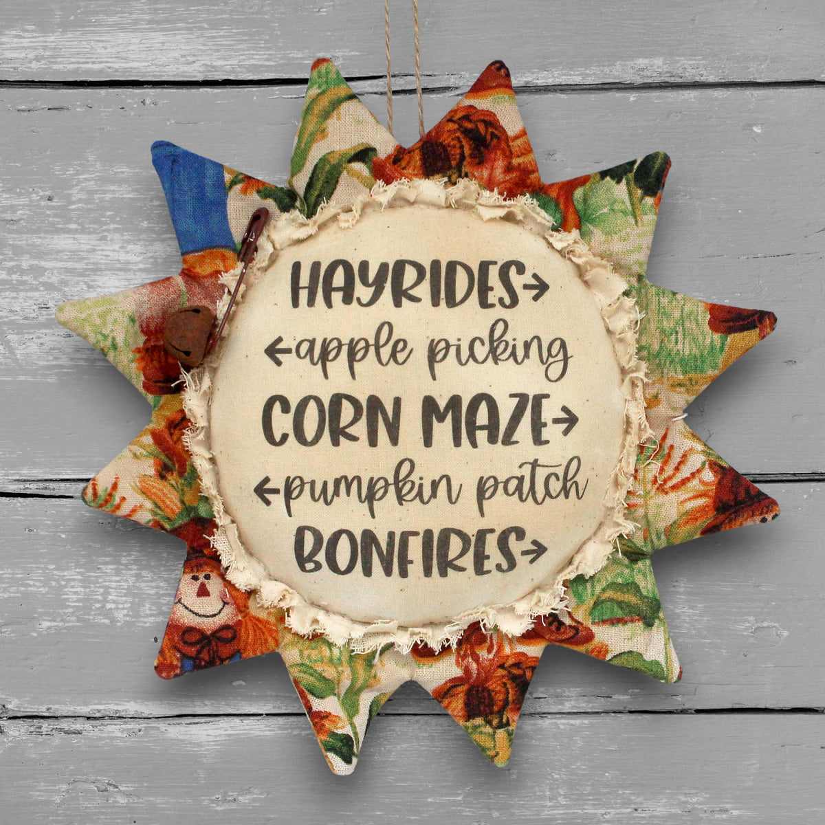 A fabric sunflower-shaped hanging ornament with a rustic cream center that reads “HAYRIDES • apple picking • CORN MAZE • pumpkin patch • BONFIRES” on a gray wood-plank background.