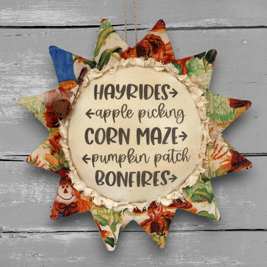 A fabric sunflower-shaped hanging ornament with a rustic cream center that reads “HAYRIDES • apple picking • CORN MAZE • pumpkin patch • BONFIRES” on a gray wood-plank background.