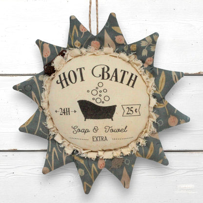 A decorative fabric sunflower ornament hanging sign with a distressed vintage 'Hot Bath' label in the center, featuring a floral patterned border and a rustic twine loop at the top, shown against a white wood plank background.